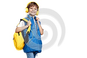 Cheerful little boy wearing headphone and yellow backpack looking to camera on white background. Back to School.