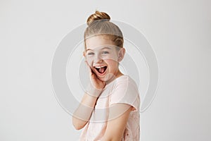 Cheerful little girl with blue eyes and bun hairstyle looks at camera with happy expression after seeing herself in