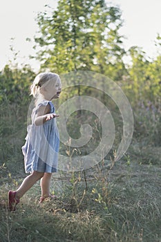 Cheerful little girl in blue sundress in park background. Cute cheerful blonde girl runs through the woods