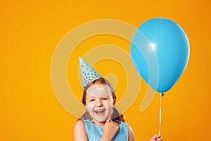Cheerful little girl celebrates birthday. The child holds a blue ball and laughs. Closeup portrait on yellow background.