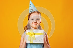 Cheerful little girl celebrates birthday. The child holds a box with a gift. A party. Closeup portrait on yellow background