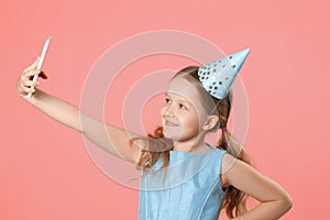 Cheerful little girl celebrates birthday. The child holds the phone, takes a selfie. Closeup portrait on pink coral background
