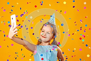 Cheerful little girl celebrates birthday. The child holds the phone, takes a selfie in the rain of confetti.