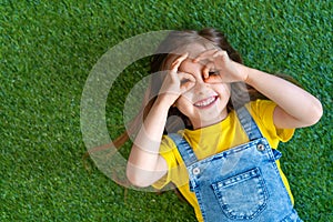 Cheerful little girl in a denim jumpsuit, lies on a green carpet, lawn