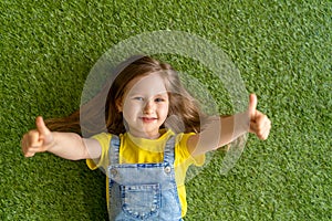 Cheerful little girl in a denim jumpsuit, lies on a green carpet, lawn