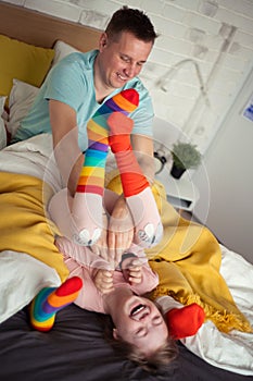 Cheerful little girl with Down syndrome lying on bed and laughing when her father is tickling her.