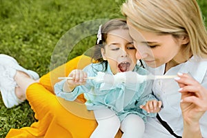 Cheerful little girl eating cotton candy with her mom, sitting on the green grass in the park.