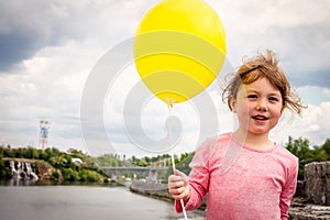 A cheerful little girl is holding a yellow ball in her hand. The child stands on the background of the river and city park. Happy