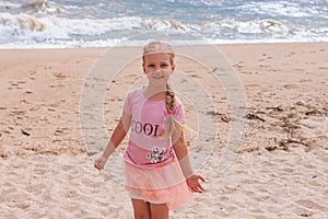 Cheerful little girl with long blonde hair in pink tulle skirt walking empty sea beach alone. Beautiful little princess