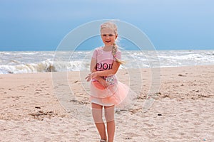 Cheerful little girl with long blonde hair in pink tulle skirt walking empty sea beach alone. Beautiful little princess