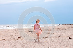 Cheerful little girl with long blonde hair in pink tulle skirt walking empty sea beach alone. Beautiful little princess