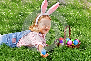 A cheerful little girl preschooler dressed in bunny ears is lying on the lawn with a basket of painted Easter eggs. Happy Easter