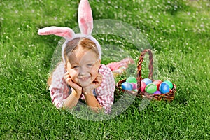 A cheerful little girl preschooler dressed in bunny ears is lying on the lawn with a basket of painted Easter eggs. Happy Easter