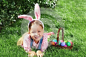 A cheerful little girl preschooler dressed in bunny ears is lying on the lawn with a basket of painted Easter eggs. Happy Easter