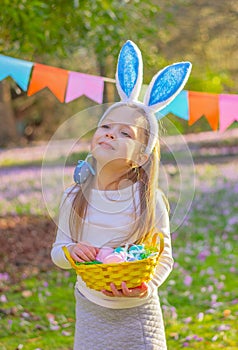 cheerful little girl with rabbit ears on her head and a basket with Easter eggs at party in garden