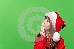 A cheerful little girl in a red knitted sweater and a Santa hat looks thoughtfully dreamily up, dreams of a gift, makes