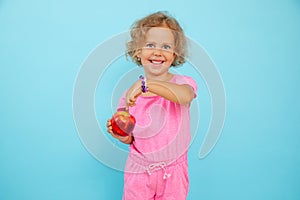 Cheerful little girl show fresh red apple on empty blue background, free copy space. Portrait of child of kindergarten