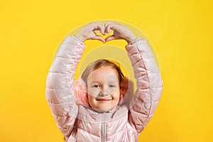 Cheerful little girl shows a heart with her fingers over her head. A child in a jacket and warm ear muffs on a yellow background.