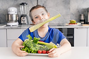 Cheerful little girl sitting with a plate of fresh vegetables and holding celery in her mouth. Cute girl having fun