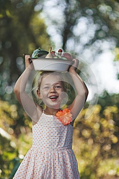 Cheerful little girl with a smile holds a plate of vegetables on her head. Rural harvesting