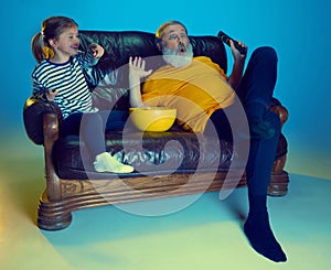 Cheerful little girl watching TV at home with her loving grandfather over blue background in neon light