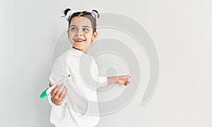 Cheerful little girl wearing white hoodie, smiling, looking at one side, and writing with greeen marker on a white board.