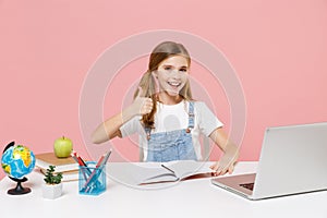 Cheerful little kid schoolgirl 12-13 years old sit study at white desk with pc laptop isolated on pastel pink background