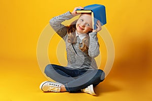 A cheerful little student girl is sitting on the floor with a book on her head. Education and school concept