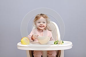 Cheerful little toddler baby girl eating healthy food while sitting in high chair looking at camera isolated over gray background