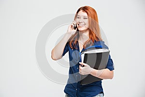 Cheerful lovely young woman with folders talking on cell phone