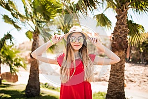 Cheerful lovely young woman in red dress and hat, sunglases walking and talking on mobile phone on summer resort