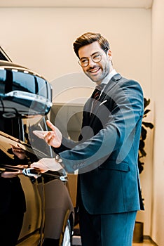 cheerful male car dealer in eyeglasses pointing at black automobile
