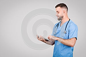 Cheerful male doctor in blue using tablet for online consult, grey background