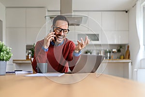 Cheerful male freelancer making business call over smart phone while working over laptop from home