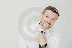 Cheerful male leader in elegant white shirt, hand on tie, smiles happily. Isolated on white background