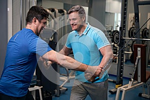 Cheerful male performing exercise on cable machine supervised by trainer