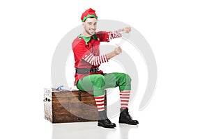 cheerful man in christmas elf costume sitting on wooden chest and holding wishlist isolated