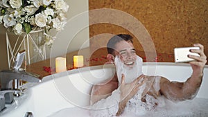 Cheerful man with foam on beard is taking selfie using smartphone in hot tub in modern spa salon. He is laughing