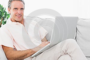 Cheerful man on his couch using laptop looking at camera