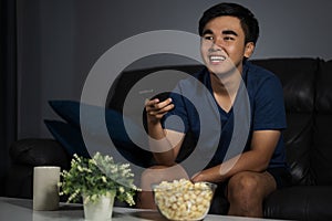 cheerful man holding remote control and watching TV while sitting on sofa at night