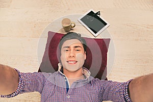 Cheerful man lying on the floor on pillow and making selfie