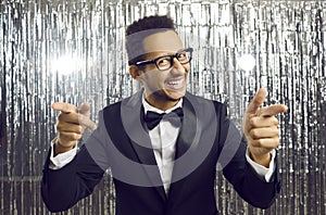 Cheerful man makes finger guns gesture directly at camera standing on shiny silver background.
