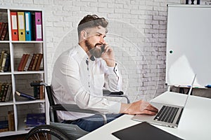Cheerful man sitting on a wheelchair, talking on a phone and using a laptop