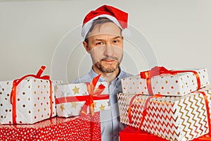 Cheerful man in t-shirt in red Santa hat holding many gift boxes standing against grey background, studio shot