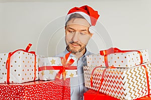 Cheerful man in t-shirt in red Santa hat holding many gift boxes standing against grey background, studio shot. Businessman is
