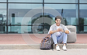 Cheerful Man Traveller Sitting Near Airport With Luggage And Using Smartphone