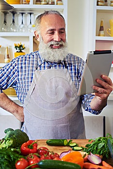 Cheerful man wearing gray apron using tablet to find a recipe