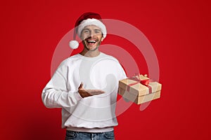 Cheerful Man Wearing Santa Hat Pointing At Gift Box In His Hand