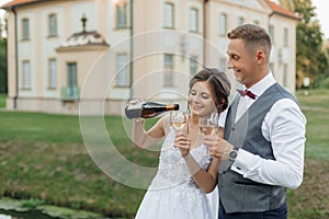 Cheerful married couple of bride in wedding dress and groom in suit pouring and drinking champagne against new house
