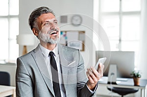 Cheerful mature businessman with smartphone standing in an office, laughing.